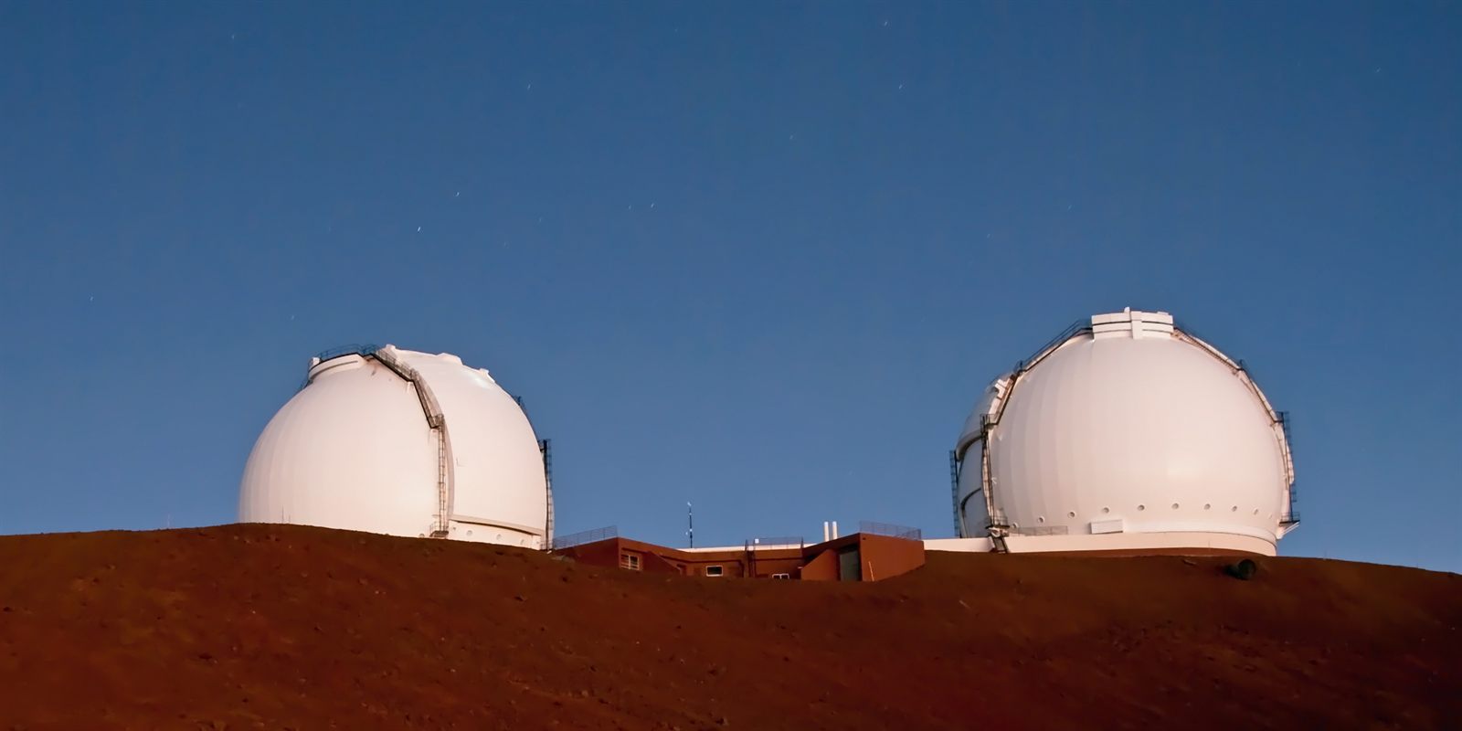 Two white observatory domes on the red cinder summit of Mauna Kea at dusk